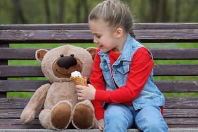 Little girl with teddy bear and ice cream on wooden bench outdoors