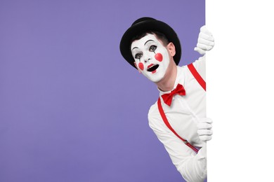 Photo of Funny mime artist peeking out of blank poster on purple background. Space for text