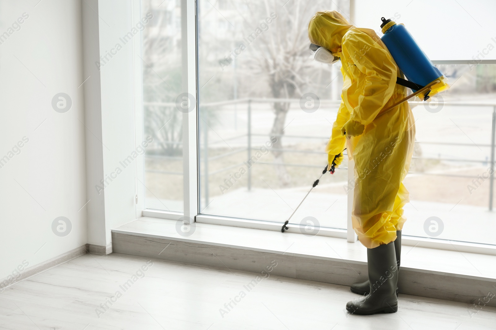 Photo of Pest control worker in protective suit spraying pesticide near window indoors