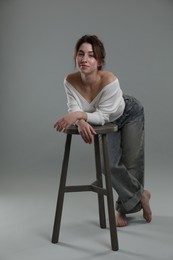 Beautiful young woman posing near stool on grey background