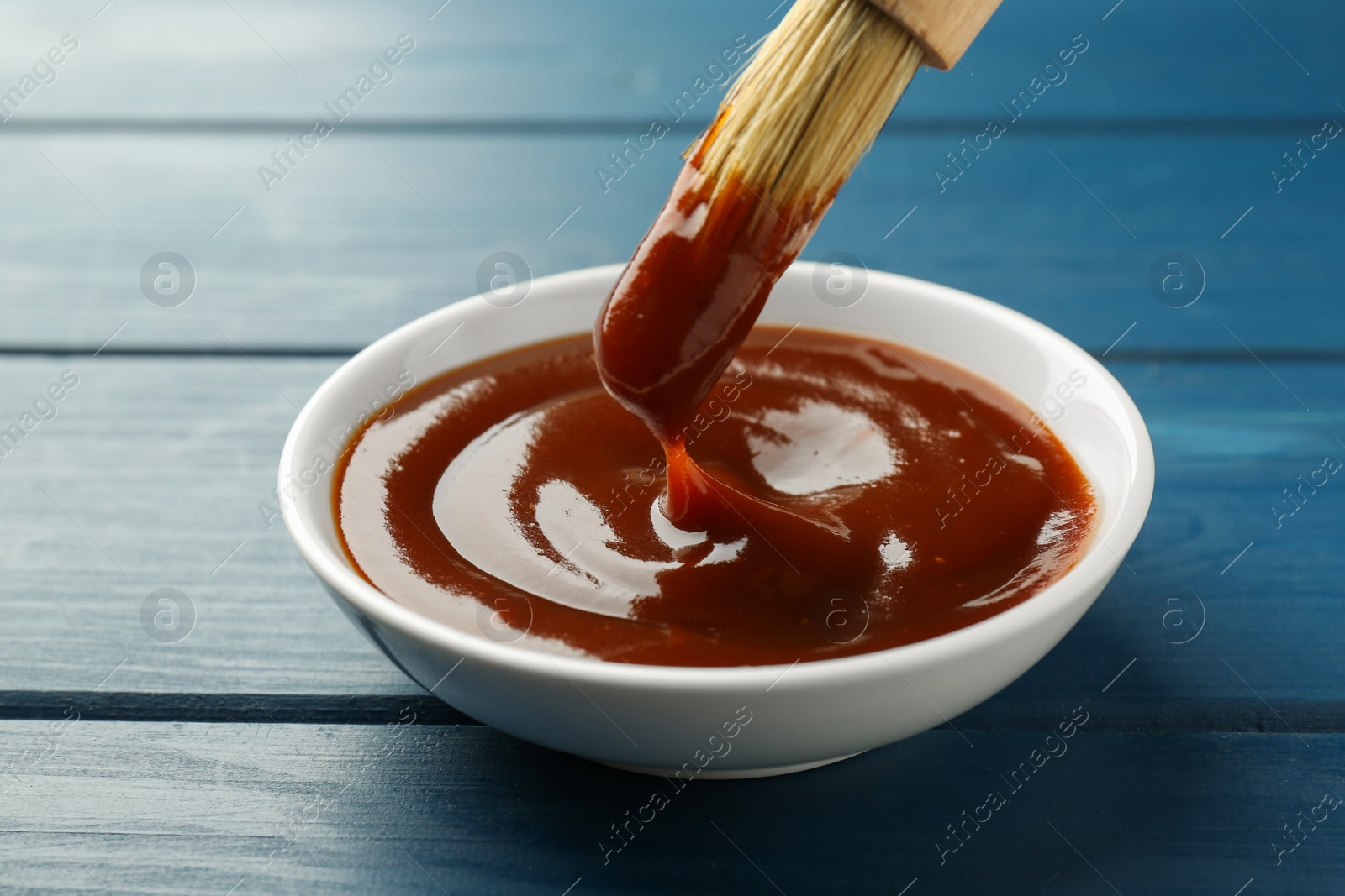 Photo of Tasty barbeque sauce in bowl and brush on blue wooden table, closeup