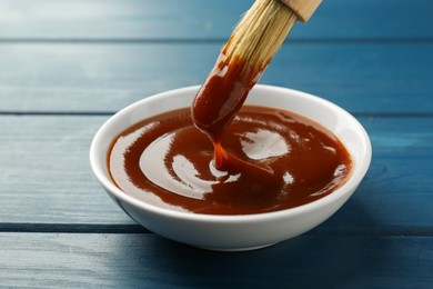 Tasty barbeque sauce in bowl and brush on blue wooden table, closeup