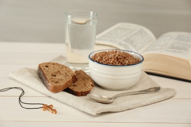 Photo of Cross, buckwheat porridge, bread, glass of water and Bible on white wooden table. Lent season