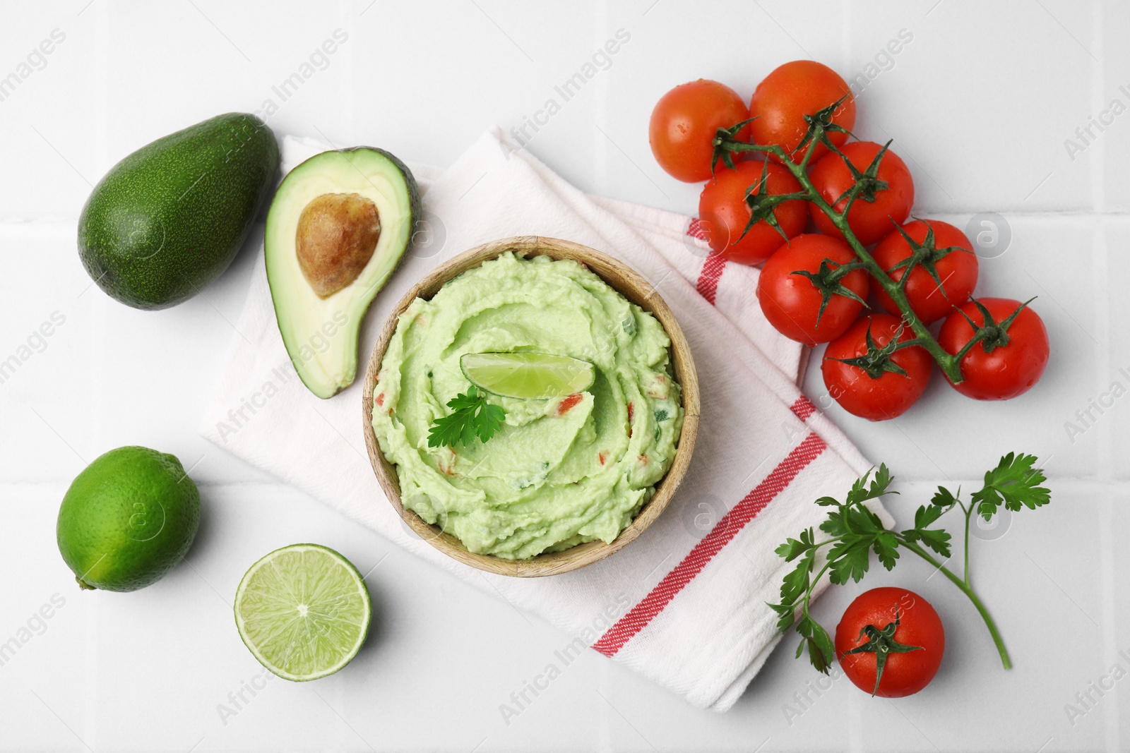 Photo of Bowl of delicious guacamole and ingredients on white tiled table, flat lay