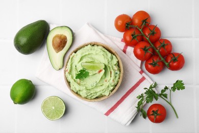 Bowl of delicious guacamole and ingredients on white tiled table, flat lay