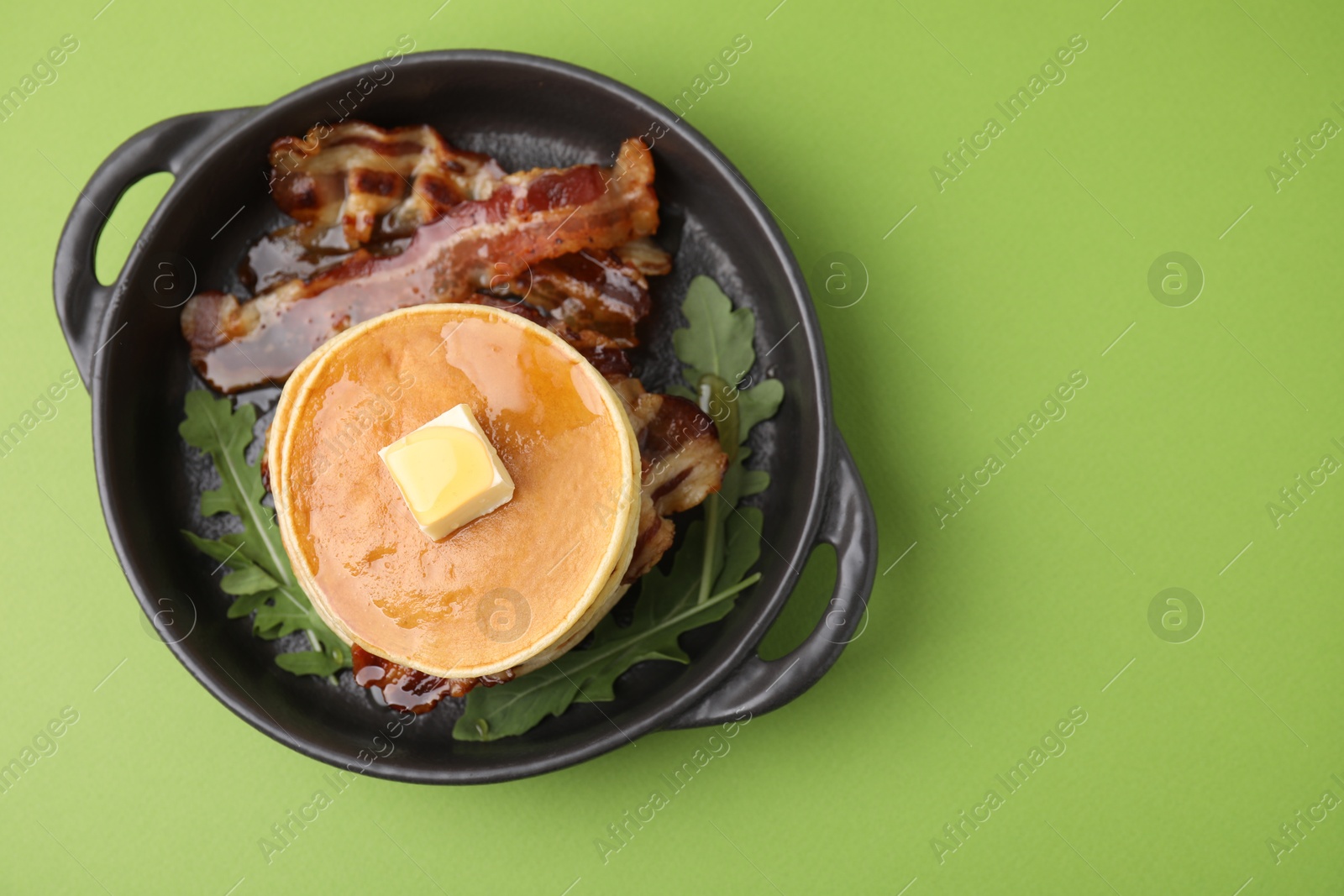 Photo of Tasty pancakes with butter, fried bacon and fresh arugula on light green background, top view. Space for text