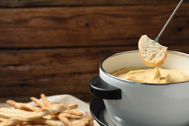 Dipping piece of bread into fondue pot with melted cheese on blurred background, closeup. Space for text