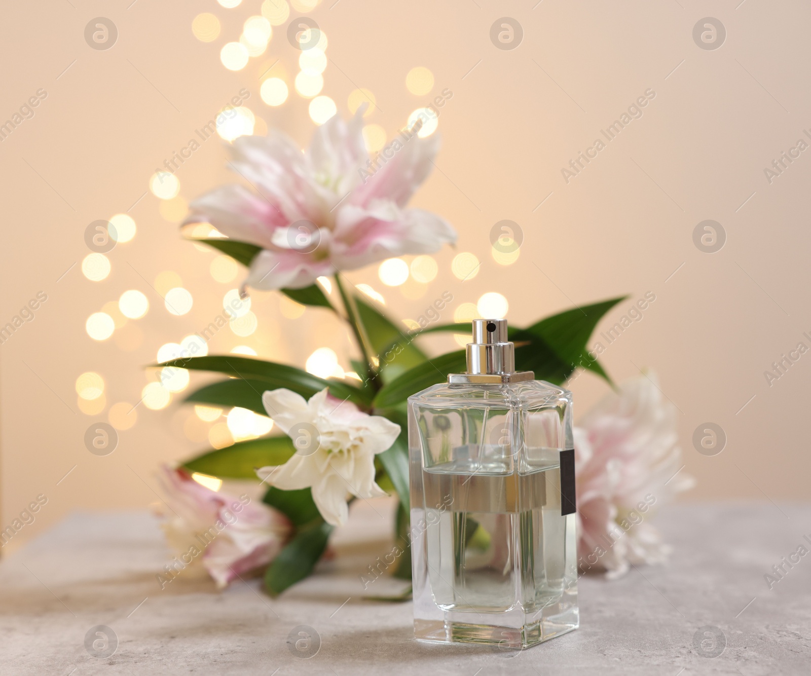 Photo of Bottle of perfume and beautiful lily flowers on table against beige background with blurred lights