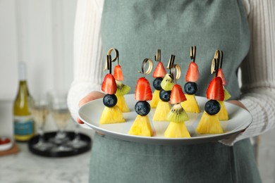 Woman holding plate of tasty canapes with pineapple, kiwi and berries indoors, closeup