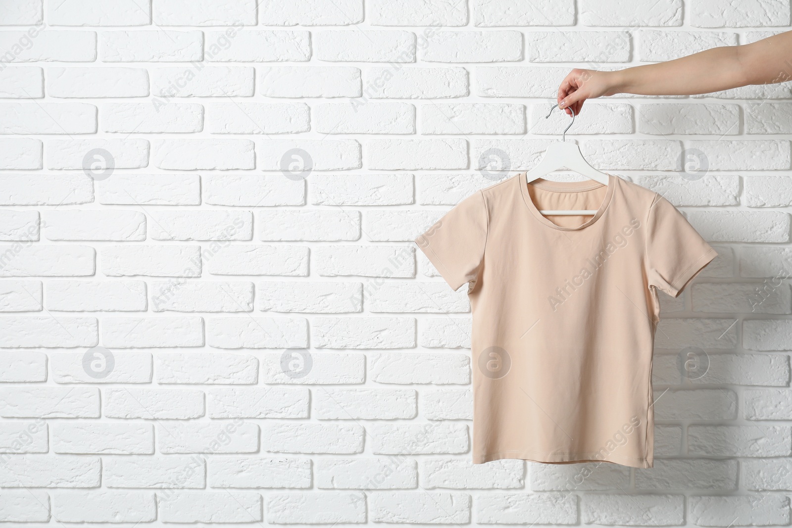 Photo of Woman holding hanger with beige T-shirt near white brick wall, closeup. Space for text