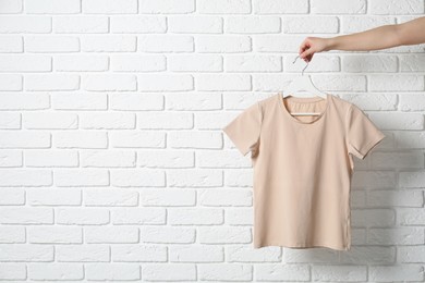 Woman holding hanger with beige T-shirt near white brick wall, closeup. Space for text