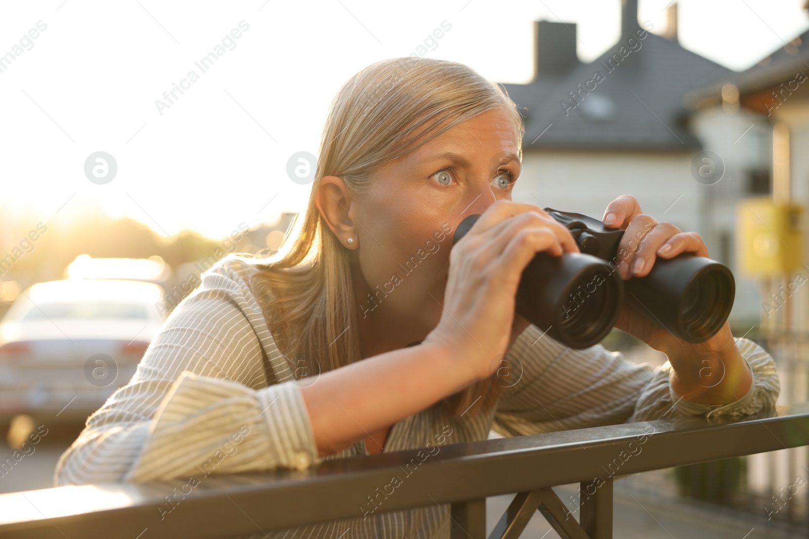 Photo of Concept of private life. Curious senior woman with binoculars spying on neighbours over fence outdoors