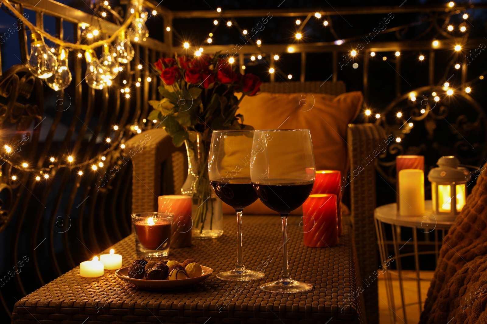 Photo of Vase with roses, glasses of wine, snacks and candles on rattan table at balcony in night