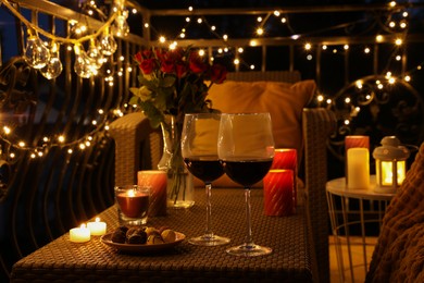 Vase with roses, glasses of wine, snacks and candles on rattan table at balcony in night