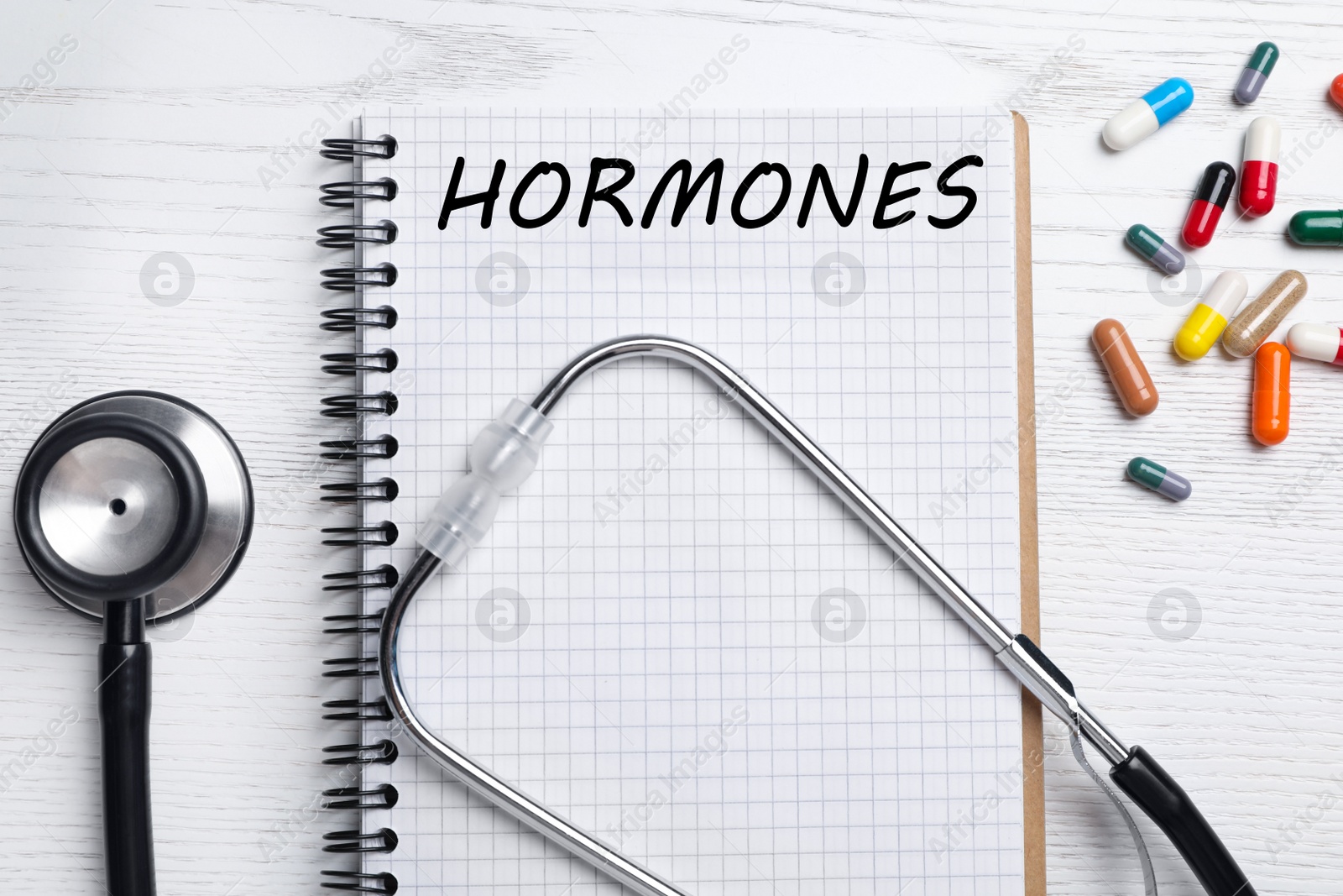 Photo of Notebook with word HORMONES, stethoscope and pills on white wooden table, flat lay