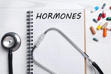 Photo of Notebook with word HORMONES, stethoscope and pills on white wooden table, flat lay