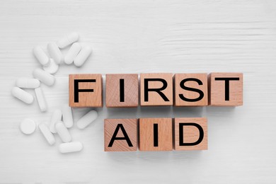 Words First Aid made of cubes and pills on white wooden table, flat lay