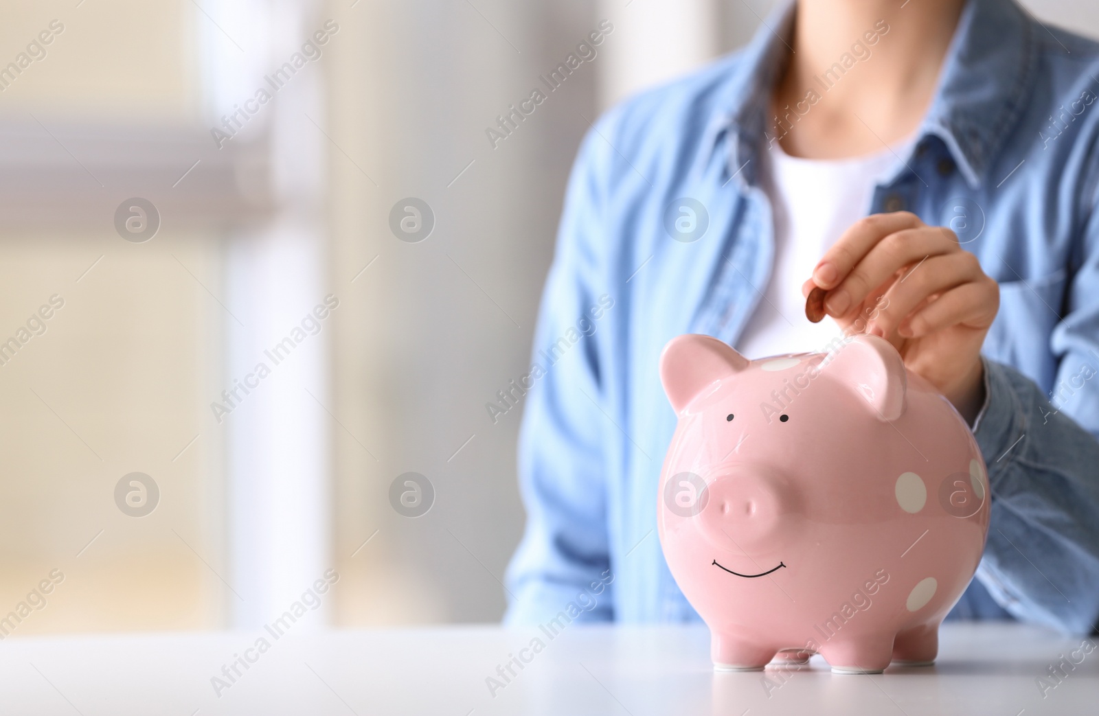 Photo of Woman putting coin into piggy bank at table indoors, closeup. Space for text