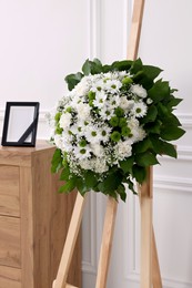 Wreath of flowers and photo frame with black ribbon on commode in room. Funeral attributes