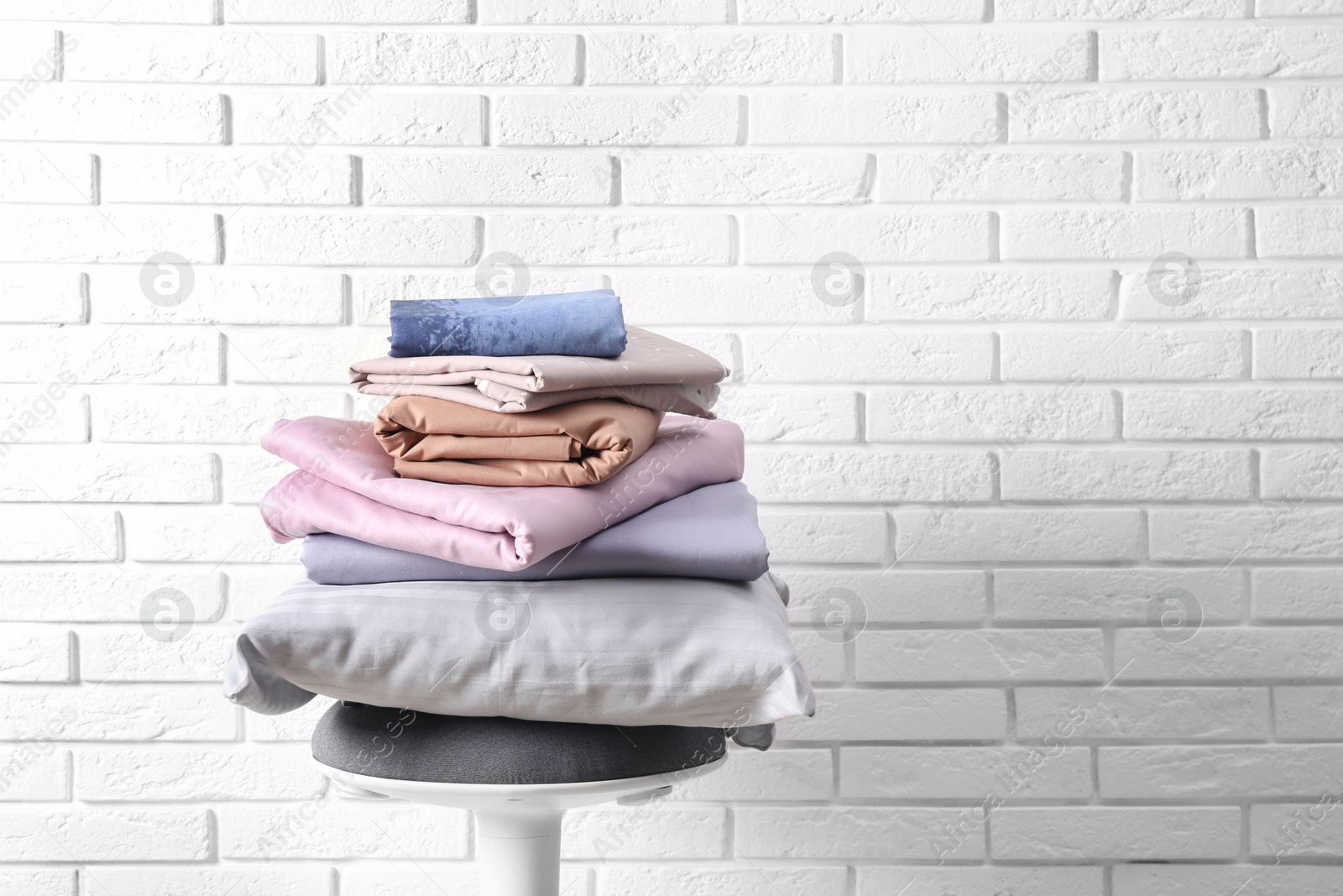 Photo of Stack of clean bed sheets and pillow on stool near white brick wall. Space for text