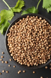 Photo of Dried coriander seeds in bowl and green leaves on dark gray textured table, flat lay