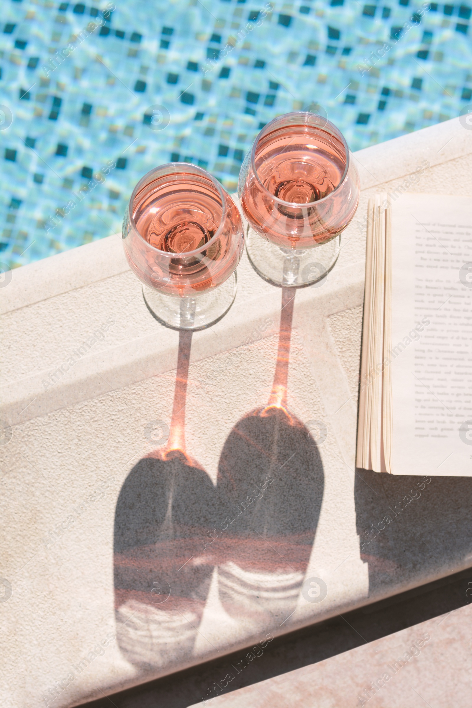 Photo of Glasses of tasty rose wine and open book on swimming pool edge, above view