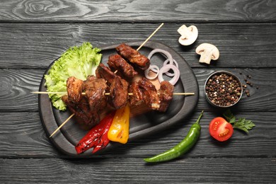 Photo of Delicious shish kebabs with vegetables and spices on black wooden table, flat lay