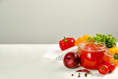 Photo of Glass jar of delicious canned lecho and fresh ingredients on white wooden table. Space for text