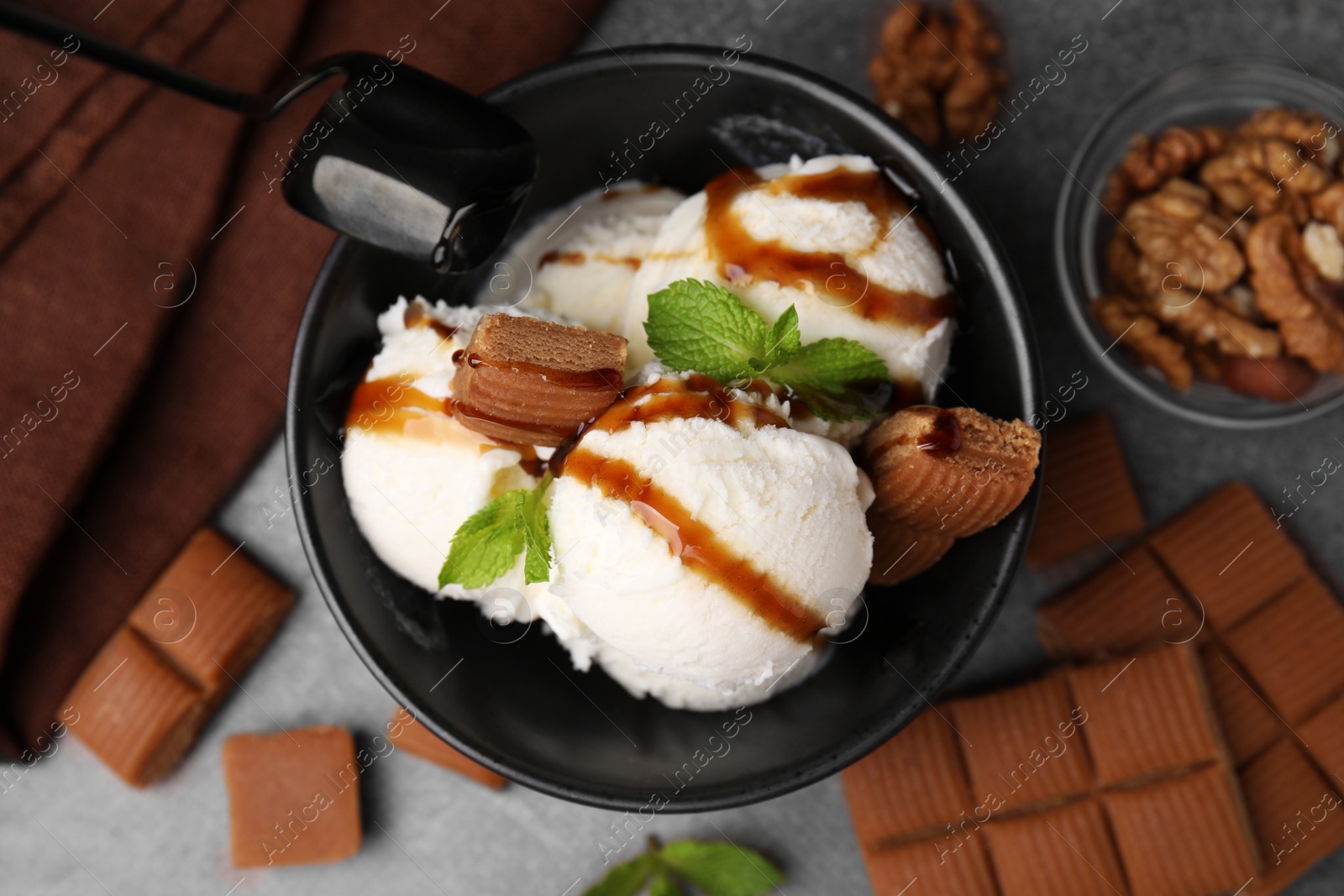 Photo of Bowl of tasty ice cream with caramel sauce, candies and mint on brown table, flat lay