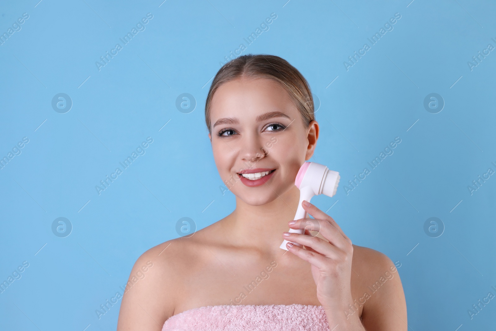 Photo of Young woman washing face with cleansing brush on light blue background. Cosmetic product