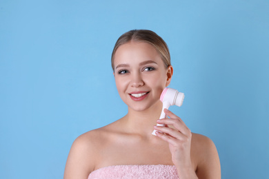 Young woman washing face with cleansing brush on light blue background. Cosmetic product
