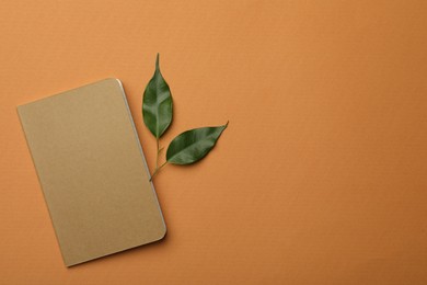 Kraft planner and green leaves on orange background, flat lay. Space for text