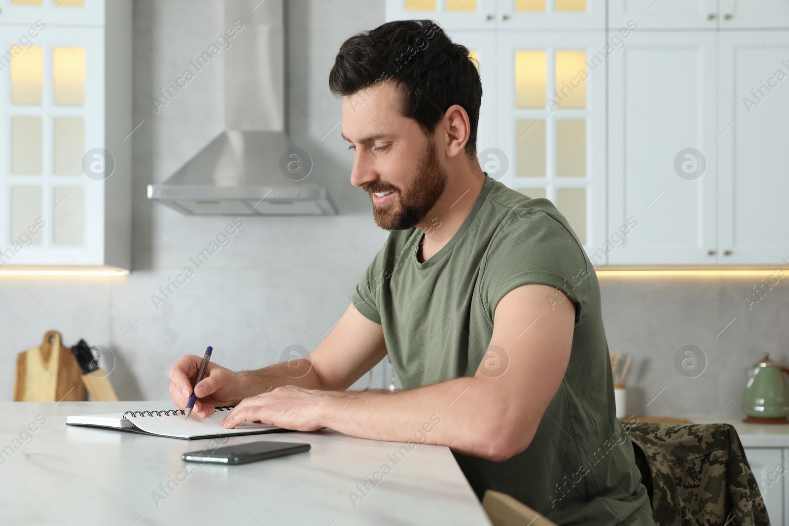 Photo of Soldier writing in notebook at white marble table in kitchen. Military service