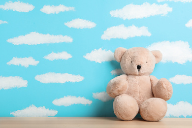 Teddy bear on wooden table near wall with painted blue sky, space for text. Baby room interior