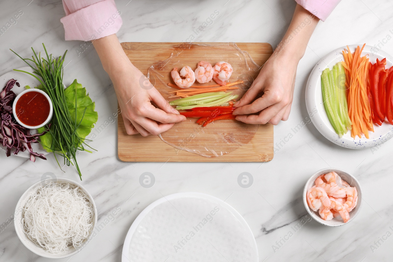 Photo of Making delicious spring rolls. Woman wrapping fresh vegetables and shrimps into rice paper at white marble table, top view