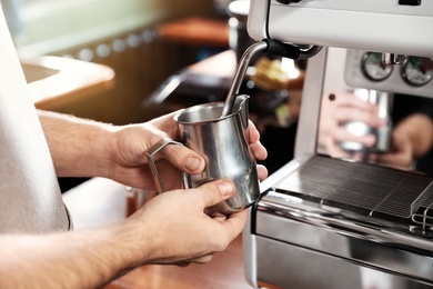 Photo of Barista frothing milk in metal pitcher with coffee machine wand at bar counter, closeup