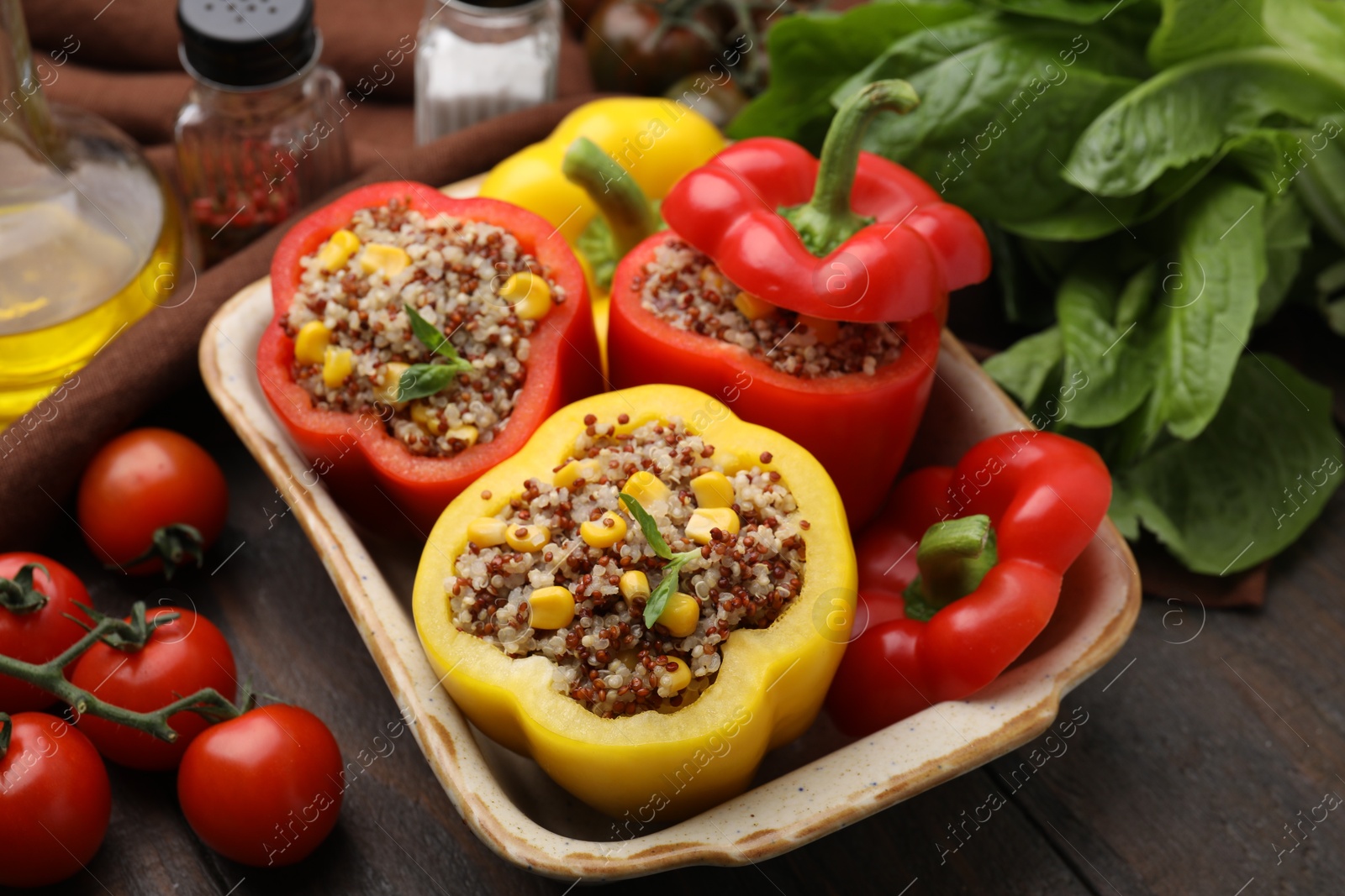 Photo of Quinoa stuffed bell peppers in baking dish, basil and tomatoes on wooden table, closeup