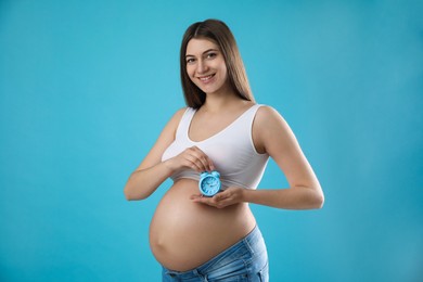Photo of Young pregnant woman holding alarm clock near her belly on light blue background. Time to give birth