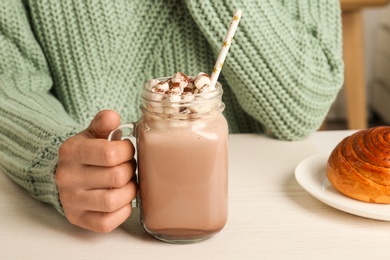Photo of Woman holding mason jar of delicious cocoa drink with marshmallows at white table, closeup