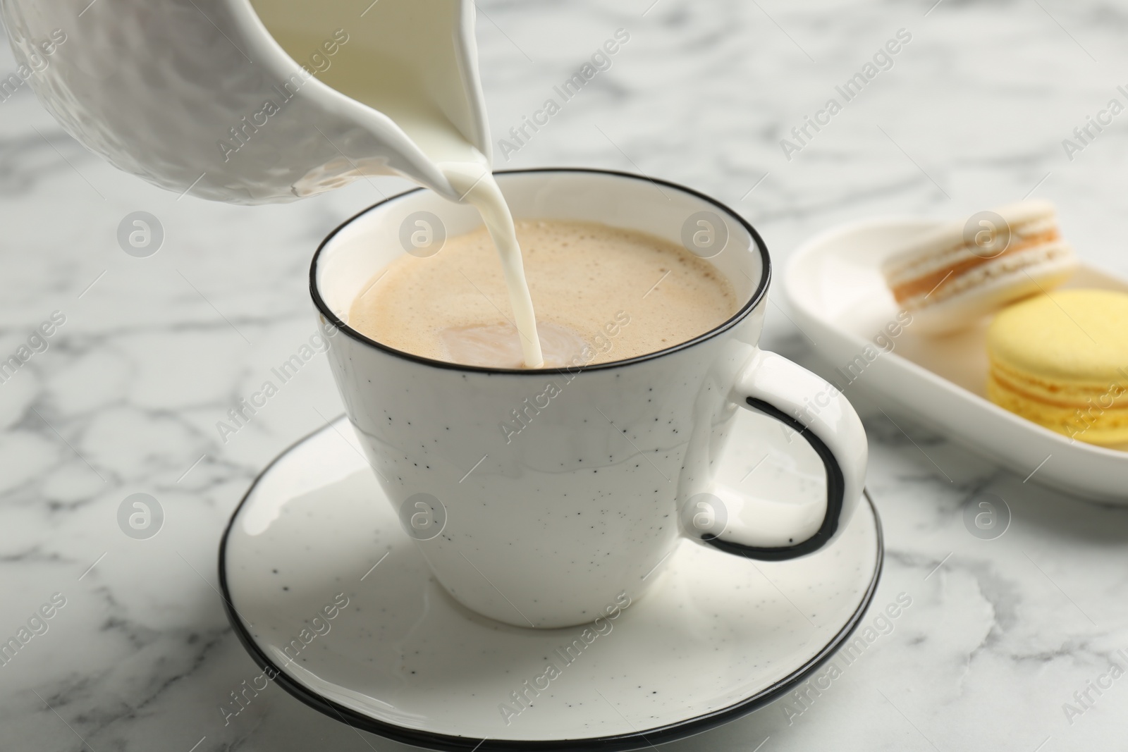 Photo of Pouring milk from pitcher into cup with coffee at white marble table, closeup