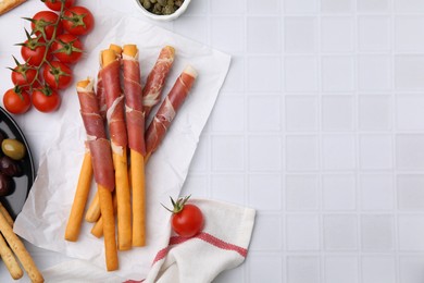 Delicious grissini sticks with prosciutto and snacks on white table, flat lay. Space for text