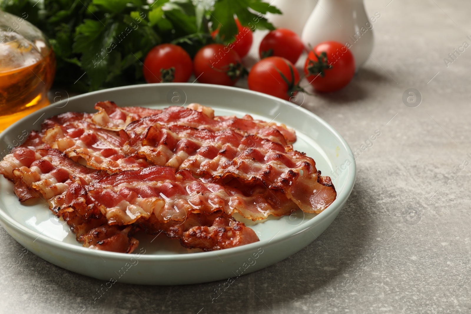 Photo of Plate with fried bacon slices and products on grey textured table, closeup