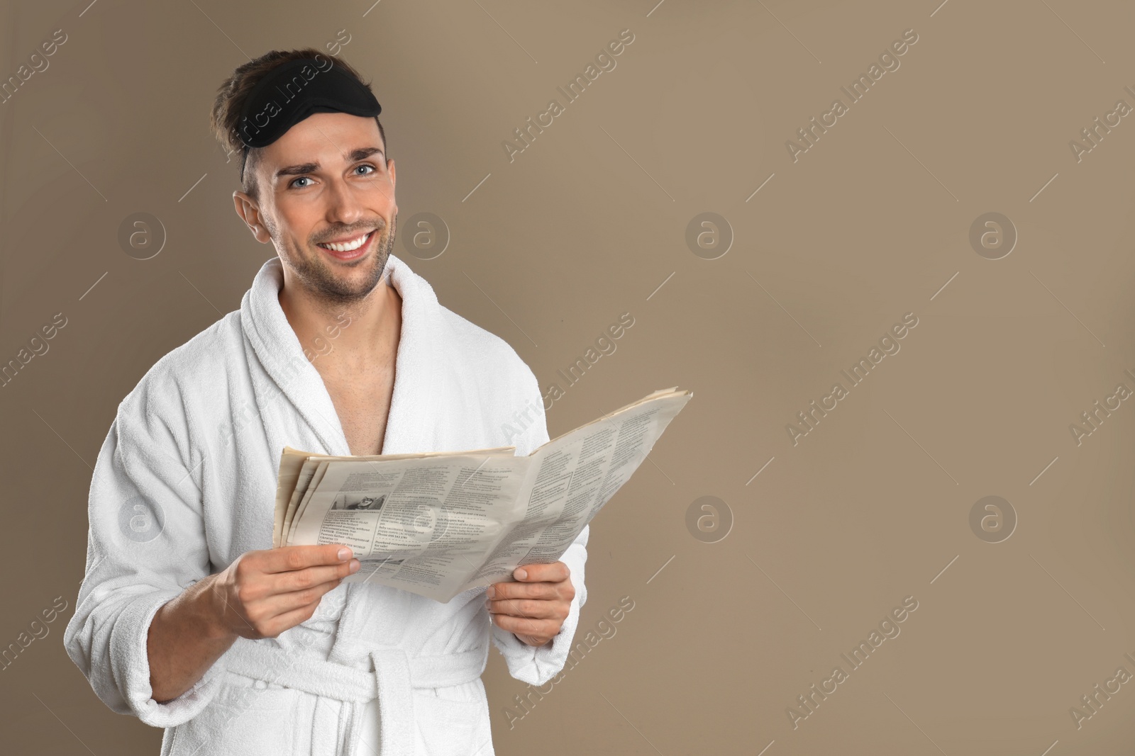 Photo of Happy young man in bathrobe and eye sleeping mask  reading newspaper on brown background. Space for text