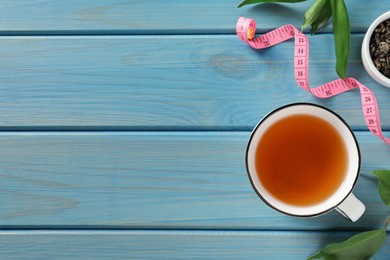 Cup of herbal diet tea and measuring tape on light blue wooden table, flat lay with space for text. Weight loss concept