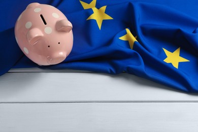 Photo of Piggy bank and European Union flag on white wooden table, above view. Space for text