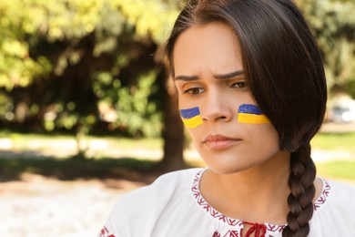 Sad young woman with drawings of Ukrainian flag on face in park, space for text