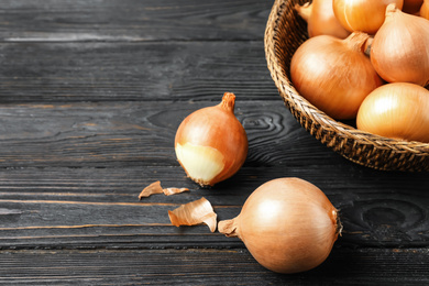 Photo of Ripe onion bulbs and basket on black wooden table, closeup