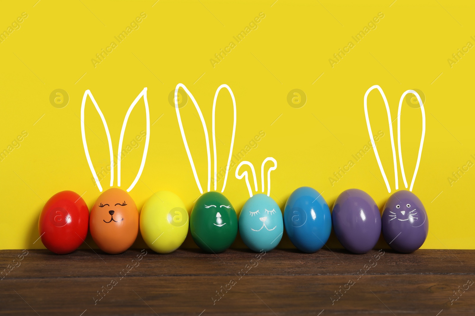 Image of Several eggs with drawn faces and ears as Easter bunnies among others on wooden table against yellow background