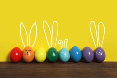 Several eggs with drawn faces and ears as Easter bunnies among others on wooden table against yellow background