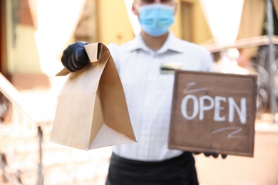 Photo of Waiter with packed takeout order and OPEN sign near restaurant, closeup. Food service during coronavirus quarantine